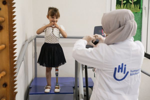 A 5-year old girl with a prosthetic leg makes a heart sign with her hands. A physiotherapist in an HI-branded white coat does the same gesture in return.
