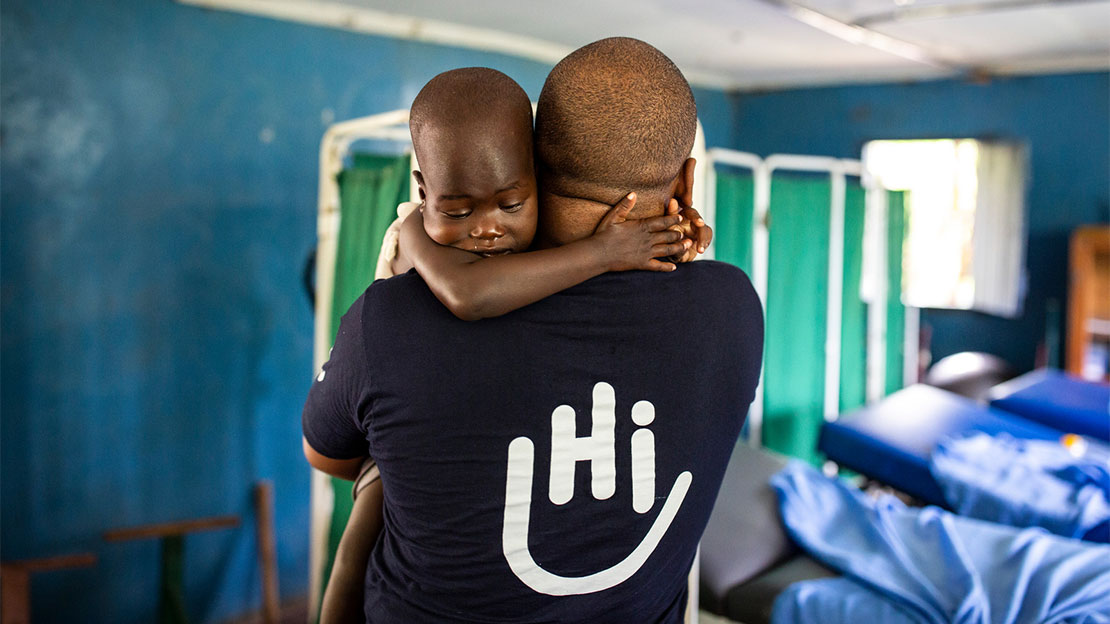 Simon Njenga, an HI occupational therapist with is back to the camera, carrying a young child who has her arms around his neck at a rehabilitation centre in Kakuma, Kenya