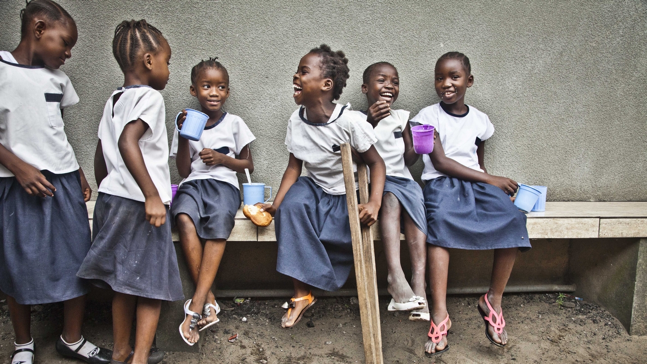 A group of young children at school sitting on a bench together, in DRC. The girl in the centre is holding a wooden crutch.