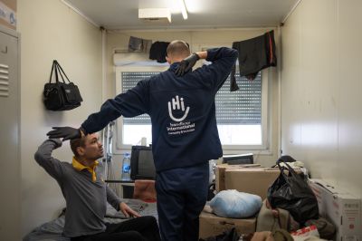 HI physiotherapist is standing up showing some rehabilitation exercices with his arm to help the beneficiary who is sitted on his bed doing the same in his bedroom