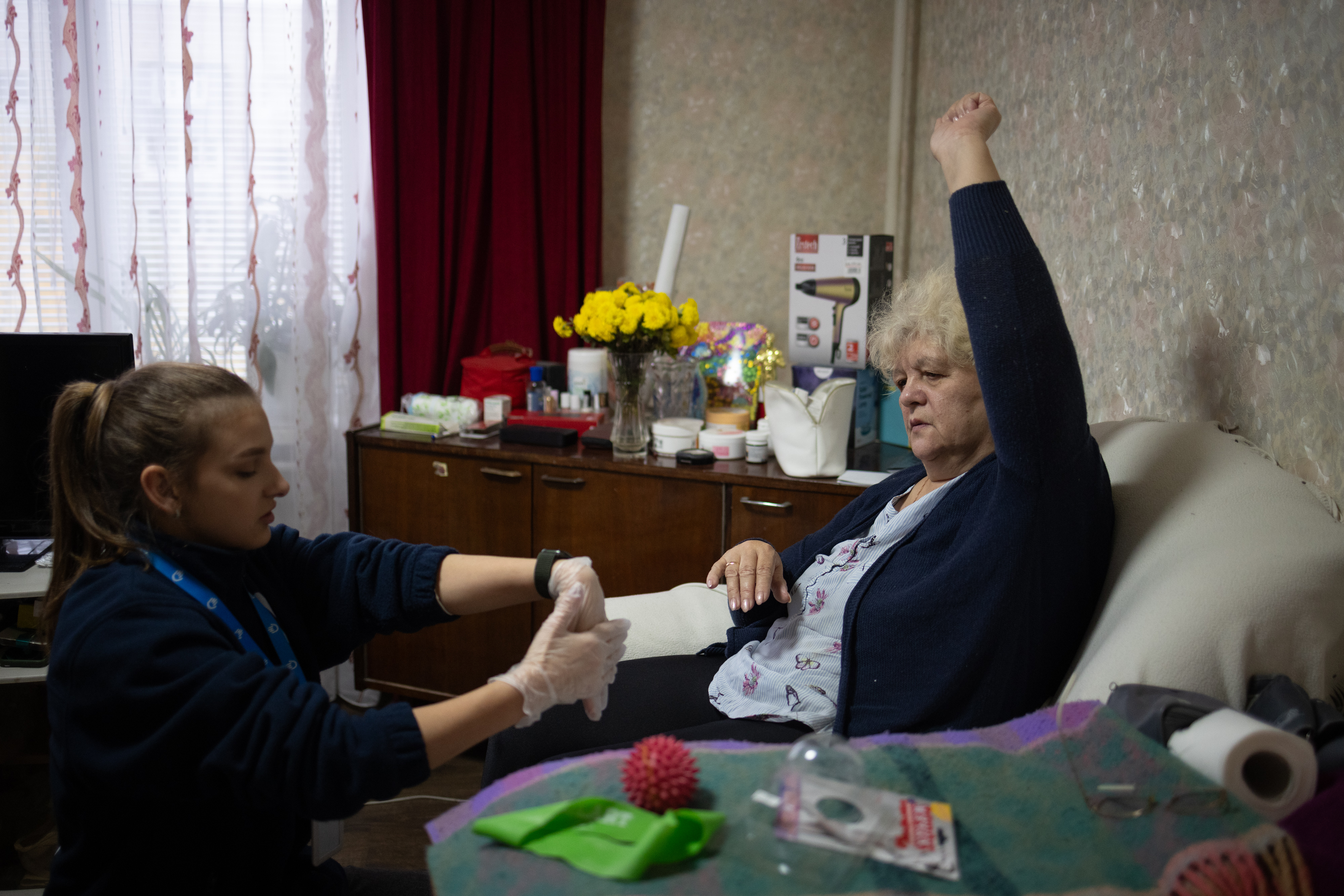 A woman sitted on a sofa making exercise with her arm raised in front of her physiotherapist Maria that is showing us rehabilitation exerices for hands
