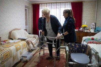 Antonina is in the center of the room walking thanks to a mobility aid with her physiotherapist we can see a sofa a table and a toilet chair