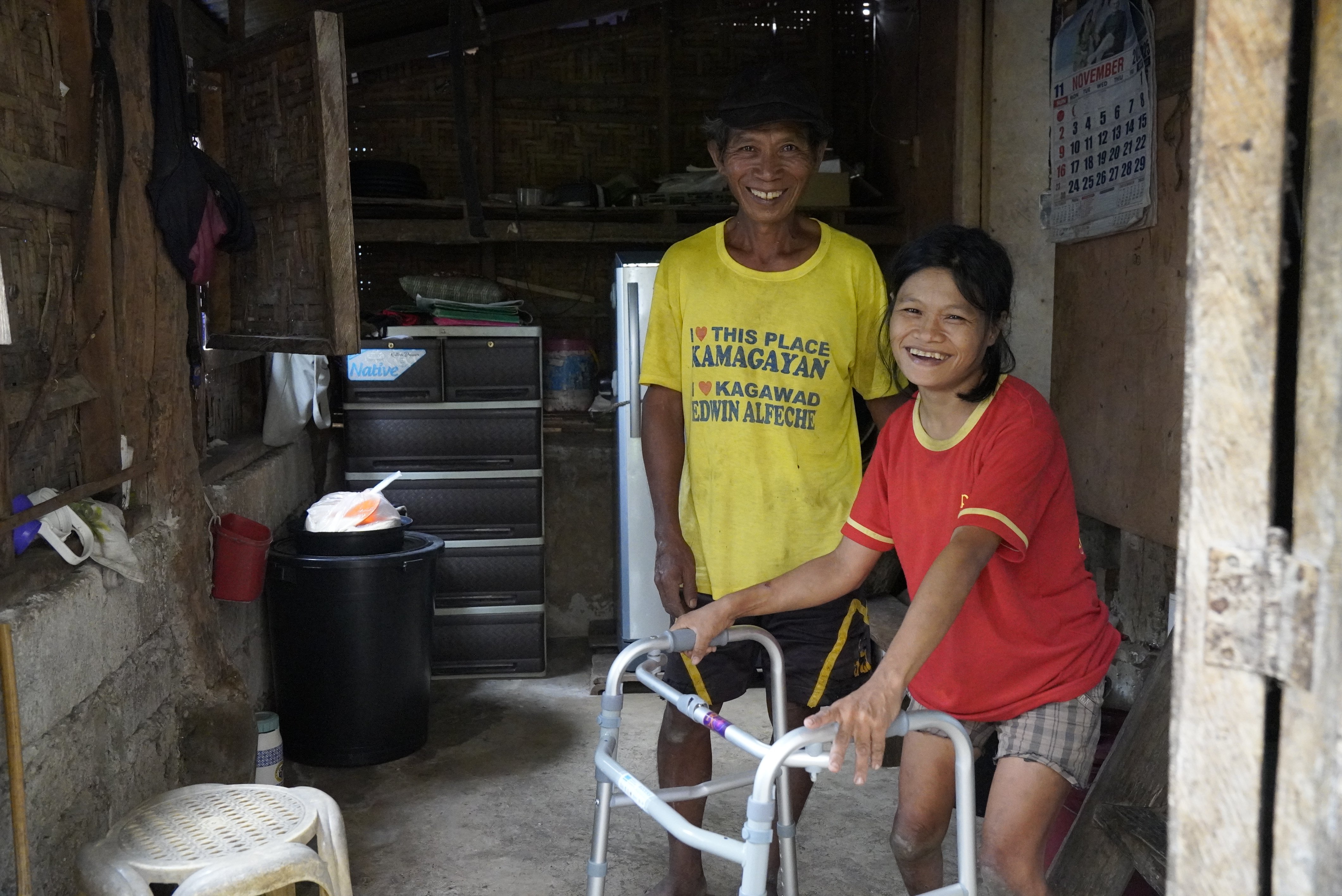 A woman with a walking frame smiles at the camera. Another man stands next to her.