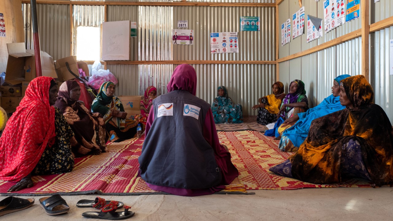 Women sit in a circle in a large room, talking to each other, accompanied by a mental health professional.