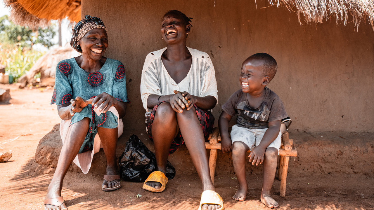 Three people are sitting in front of a hut, laughing out loud.