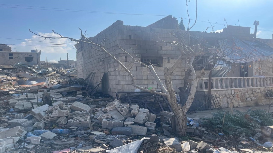 The photo shows a partially destroyed brick building, whose roof and some walls appear to have collapsed. In the foreground, a leafless tree stands amid a large pile of rubble: broken concrete blocks, pieces of wood, metal fragments, and various scattered objects. To the right, another structure, built of stone and covered with damaged sheet metal, also appears to have been damaged. The ground is completely littered with rubble, and in the background, other damaged buildings can be seen under a clear sky.  