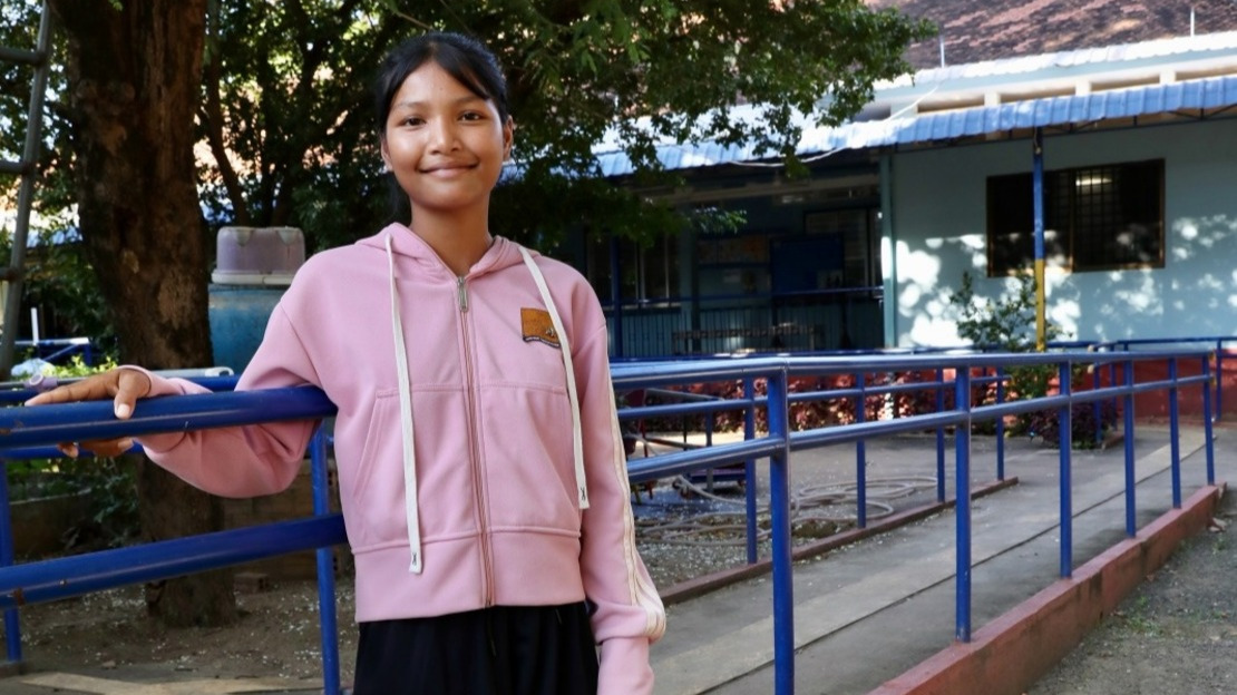 Portrait of Srey Neang leaning against a physiotherapy exercise bar in the courtyard of the rehabilitation centre. Behind her, a large tree and an exercise ramp can be seen.