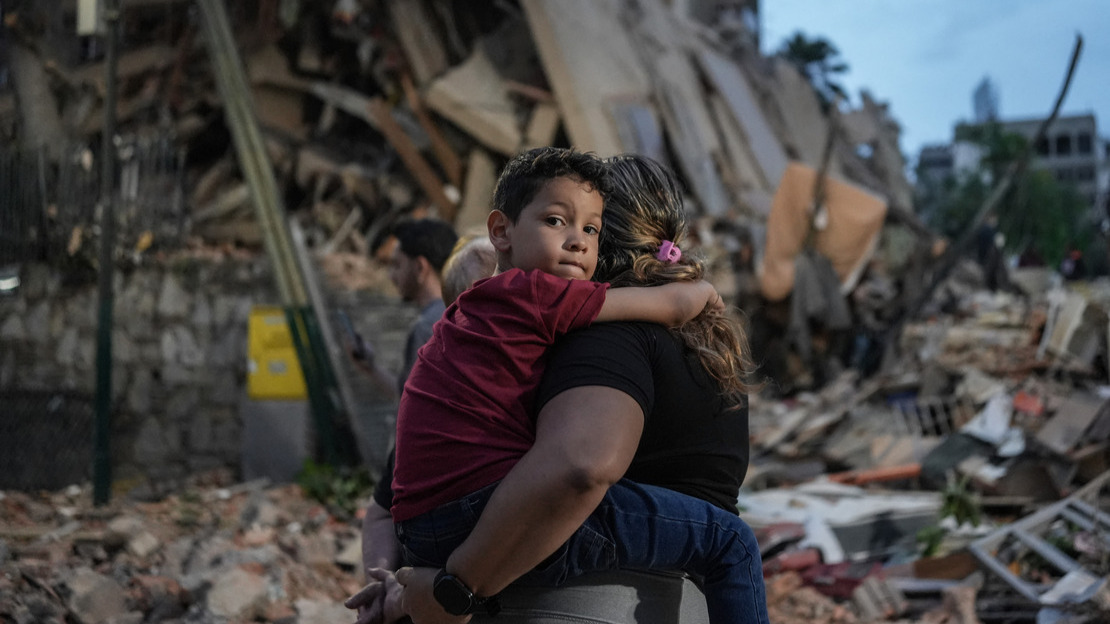 In the foreground, a woman, seen from behind, is holding a child in her arms; the child is staring intently at the camera. Behind them, collapsed buildings can be seen.