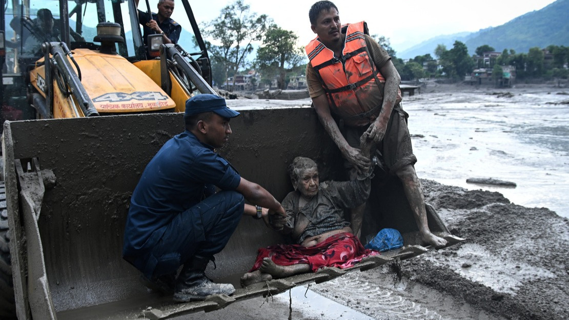 Two men are supporting a disabled woman who is half-reclining in the bucket of a backhoe loader. They are all covered in mud, and the landscape around them is flooded.