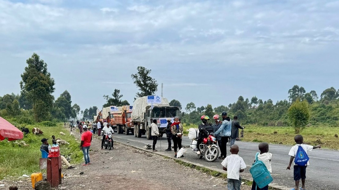 A line of trucks displaying the HI and EU logos moves along a road. At the side of the road, many people on motorcycles or on foot carrying packages or watching the trucks go by.