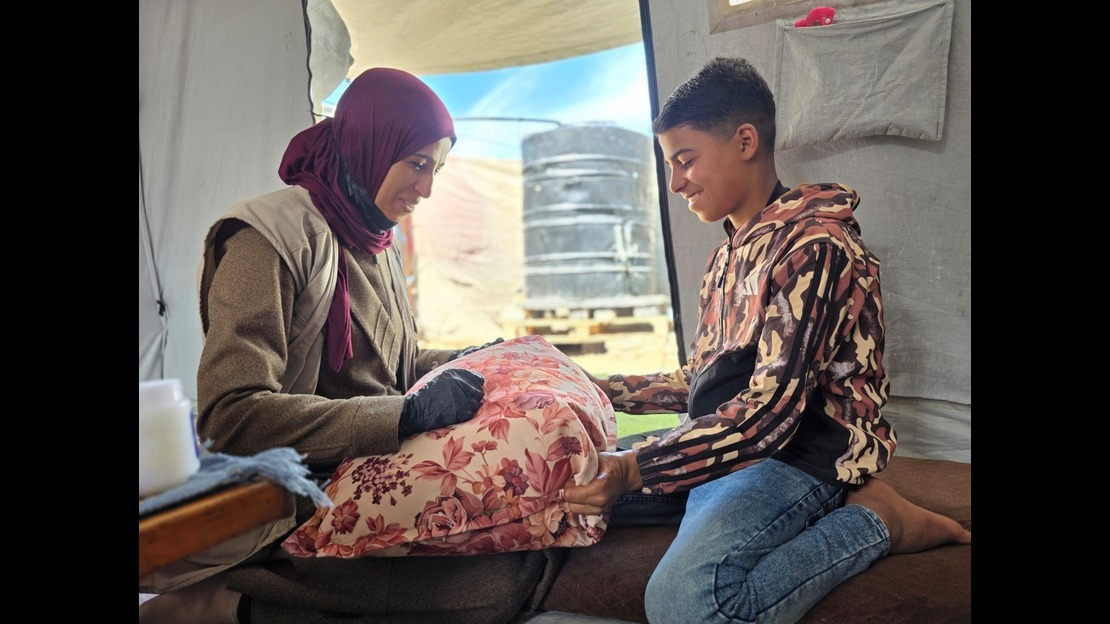 Inside a canvas shelter, two people are sitting facing each other on cushions. An adult is wearing layered clothing and a scarf covering their hair, and is holding a floral-patterned cushion on their lap. Across from them, a younger person, kneeling on a rug, wears a patterned hoodie and jeans, and is also holding the cushion with their hands. In the background, an opening reveals a blue sky and hanging fabrics. Natural light illuminates the scene through the opening. 