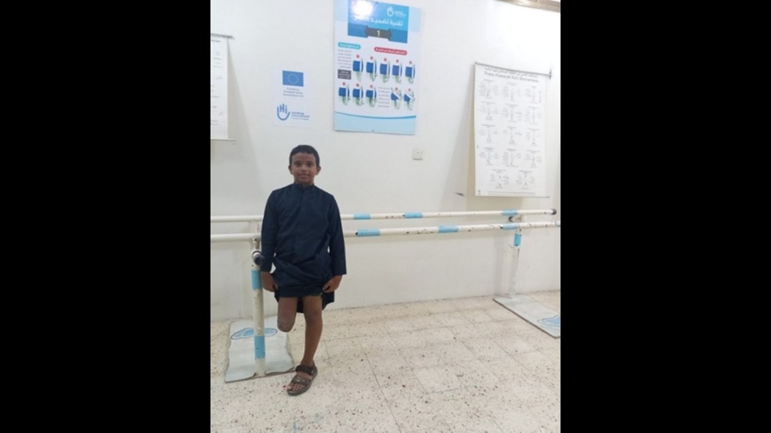 A young boy is standing in a physical therapy room, leaning slightly on white and blue parallel bars anchored to the floor, which are used for walking practice. The room is brightly lit, with medical and educational posters hanging on the wall in the background.