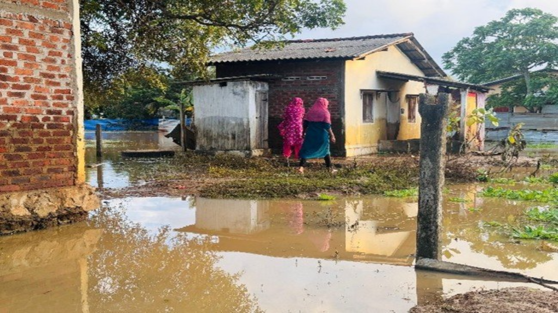 Two women dressed in colorful clothes walk through a flooded area, near brick and concrete houses. The ground is covered with muddy water.