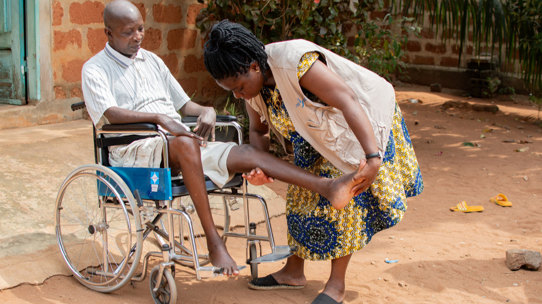 A man sitting in a wheelchair watches the movement of a woman standing next to him, who is extending his left leg.
