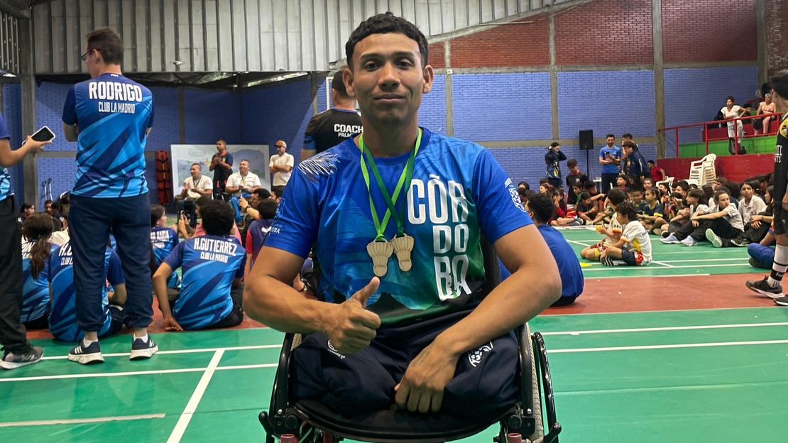 A young man in a wheelchair is wearing a sports shirt and has two medals hanging round his neck. Behind him, an indoor stadium can be made out.