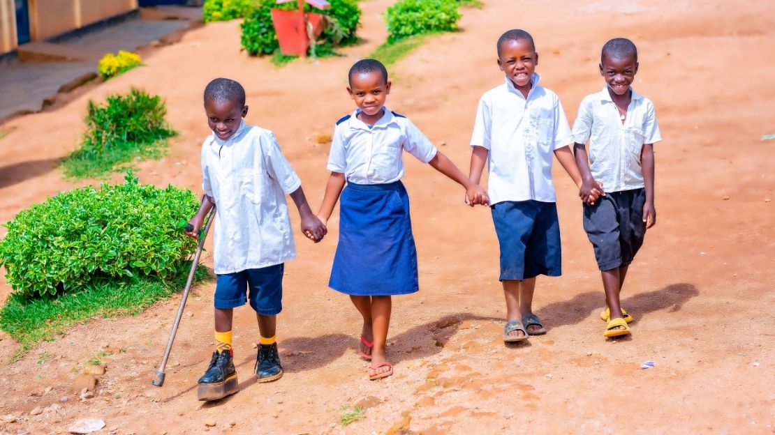 Four children in school uniforms are walking hand in hand. The boy on the left in the photo is wearing an orthopedic shoe and using a crutch.