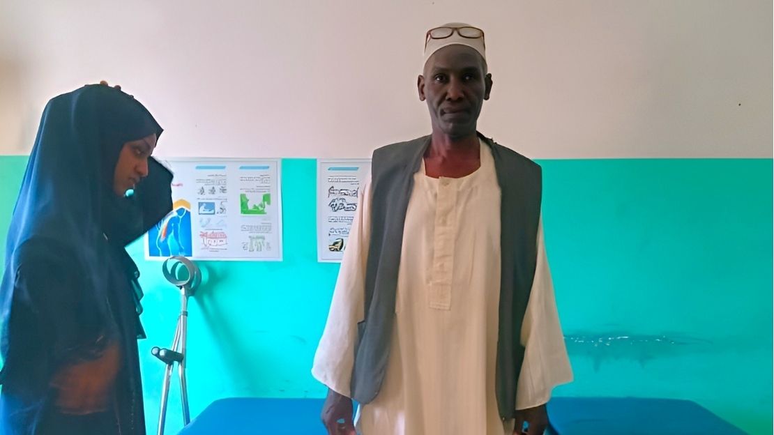 A man is standing and looking at the camera. In the background, a few crutches are leaning against the wall.