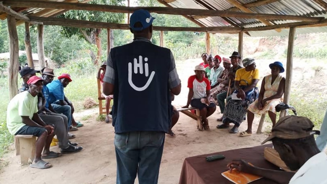 Under a sheet-metal shelter, a man stands with his back to us, wearing a T-shirt with the HI logo. Facing and surrounding him, seated on benches, men and women listen to him.