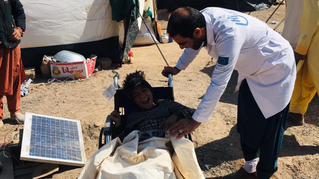 A HI physiotherapist stands next to a child in a wheelchair near the shelters. 