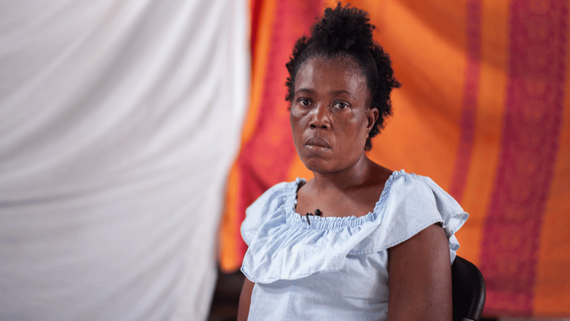 A close-up portrait of a Black woman sitting in a chair. She looks at the camera with a contemplative and slightly sad expression.