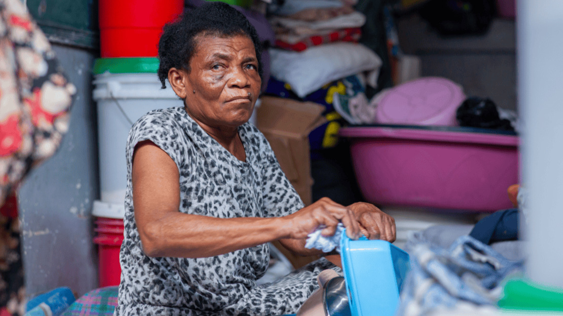 A woman is sitting and cleaning a plastic container with a cloth. Behind her, one can make out a pile of buckets, cloths, cardboard boxes, and so on.