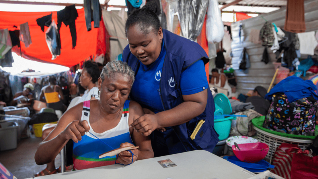 A seated woman holds a wooden object through which she threads a lace. A woman wearing a HI jacket leans over her. Behind them, we can make out tarpaulins, metal sheets, clothes drying and people sitting down.
