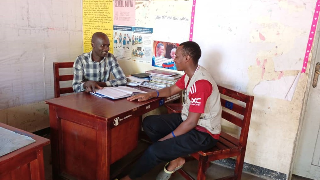 Two adults are talking in a classroom, on either side of a desk.