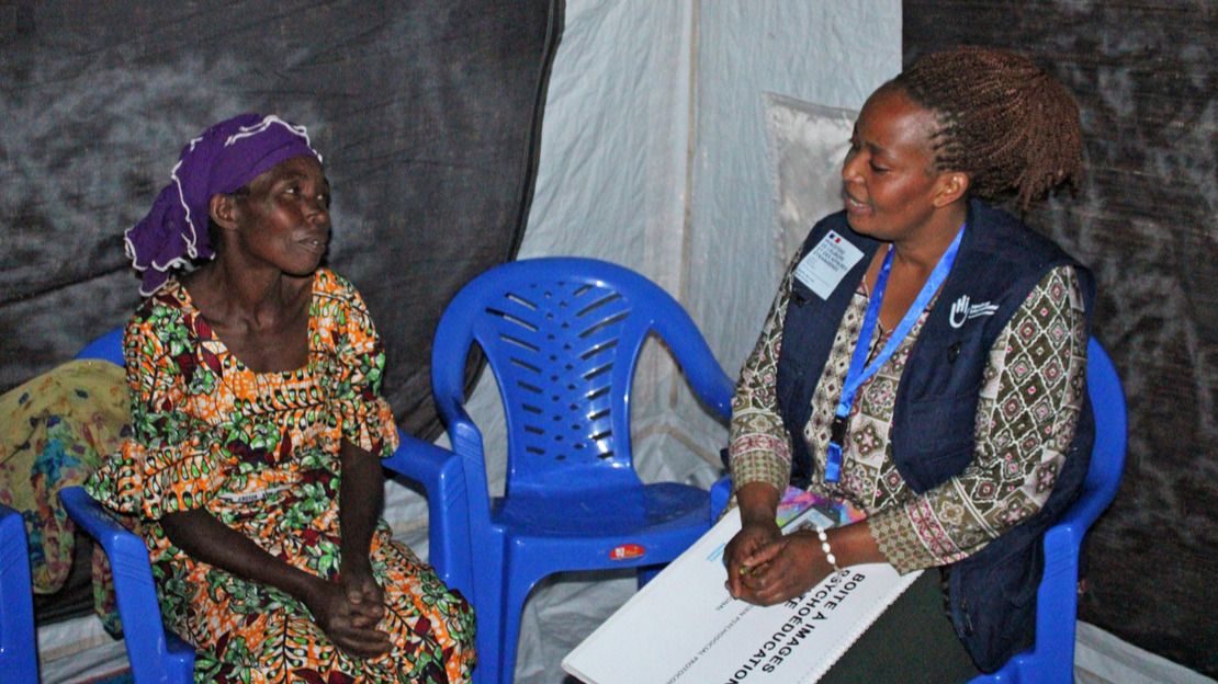 Two women are sitting on chairs in a tent. They are looking at each other and talking. The woman on the right is wearing a jacket with HI printed on it and is holding a notebook entitled “psychoeducation image box” on her lap.