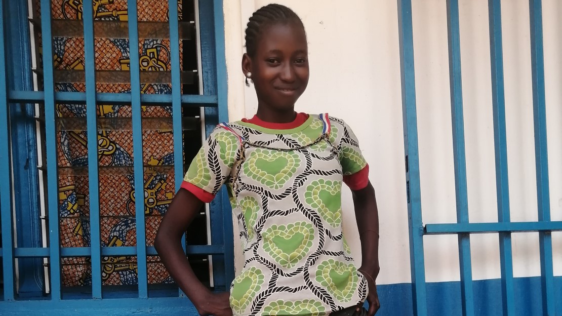 A teenager is standing in front of the HI rehabilitation room, she's smiling and shows off her prosthesis.