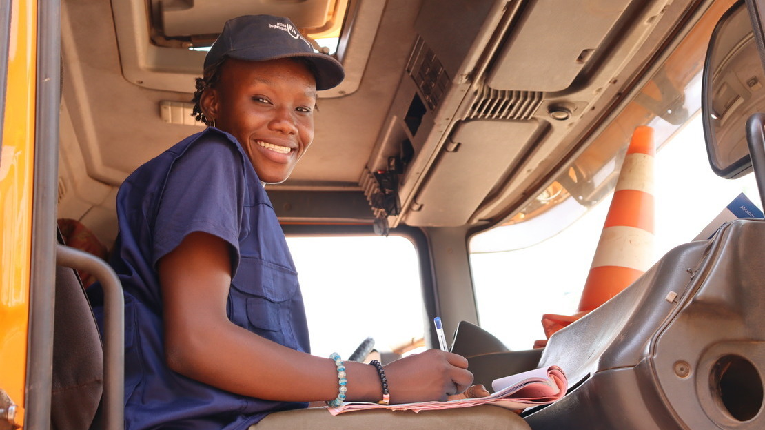 A young black woman sits in the cab of a truck. She's holding a pen and papers and looking down at the camera with a smile.
