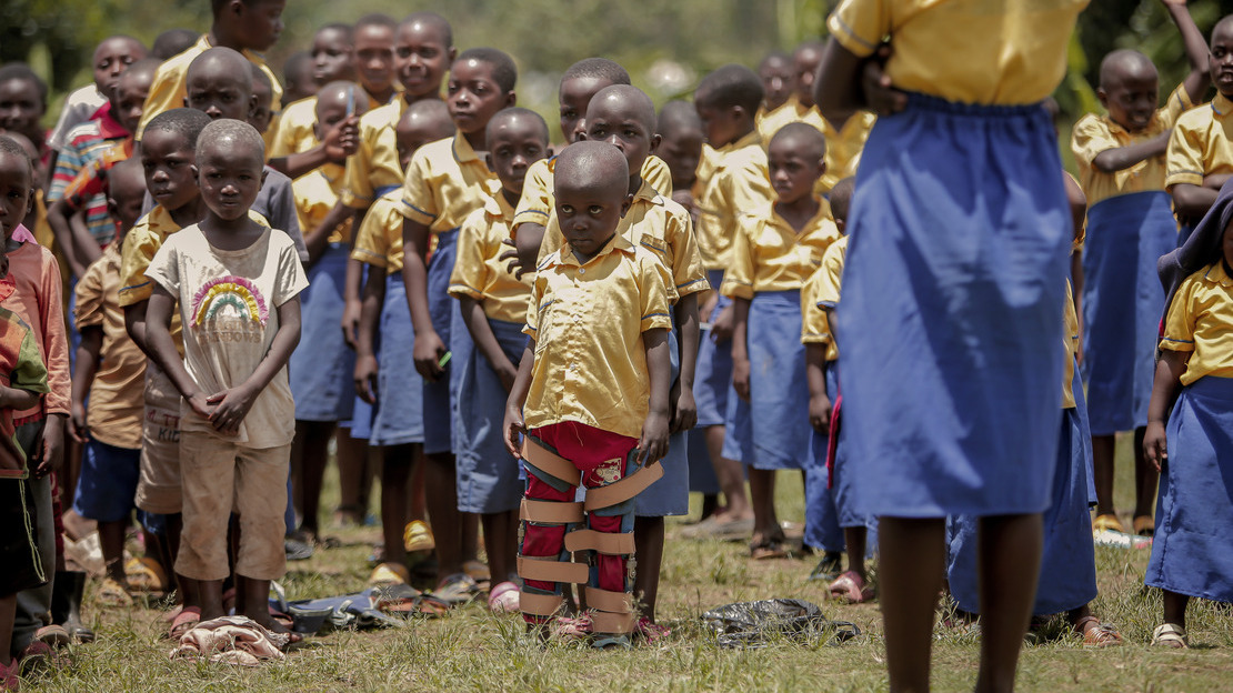 Children in school uniforms stand single file in front of an adult seen from behind. In the front row, a little girl wearing braces on both legs. 