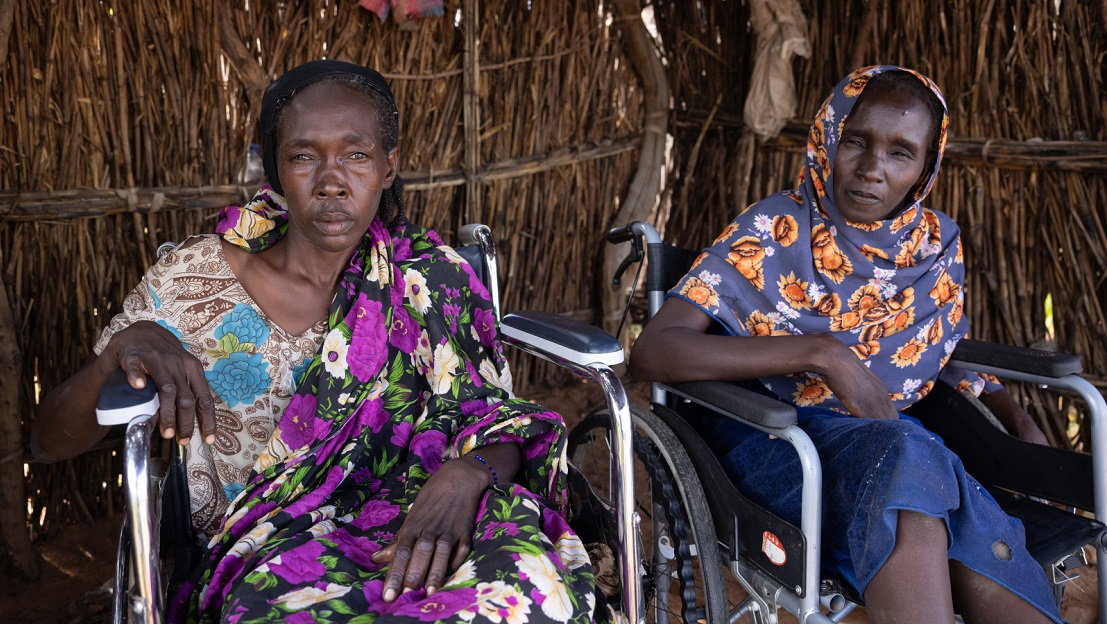 Kaltouma and Hassaneih are side by side, each in a wheelchair. They are sheltered in a wooden house, looking at the lens. Katlouma, on the left, wears a dress with purple and white flowers, while Hassaneih, on the right, wears a blue scarf with orange flowers. 