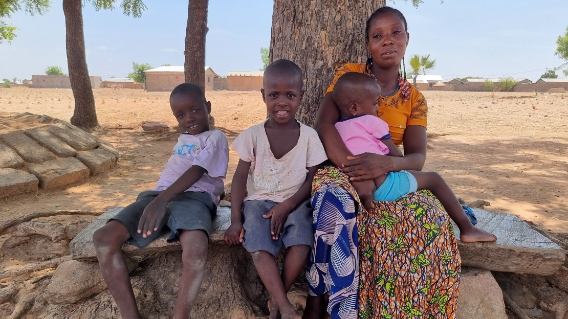 The image shows a woman and two children sitting side by side on a stone bench at the foot of a large tree. The woman is holding a child in her arms. The background shows a dry rural landscape with a few scattered trees and houses visible in the distance.
