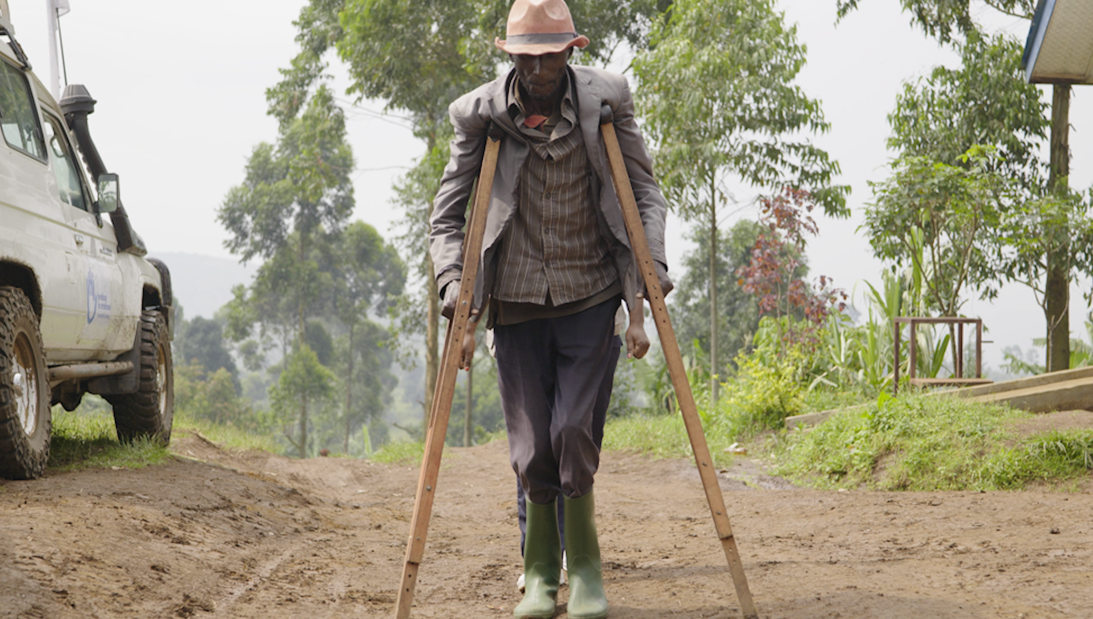 A person is walking along a dirt path, leaning on two long wooden crutches held on either side of their body. The person is wearing a jacket, a striped shirt, pants, and green rubber boots, as well as a hat. The path winds through a landscape lined with trees and plants. On the left side of the image, a white vehicle is partially visible, parked along the path. In the background, another person is visible behind the person.
