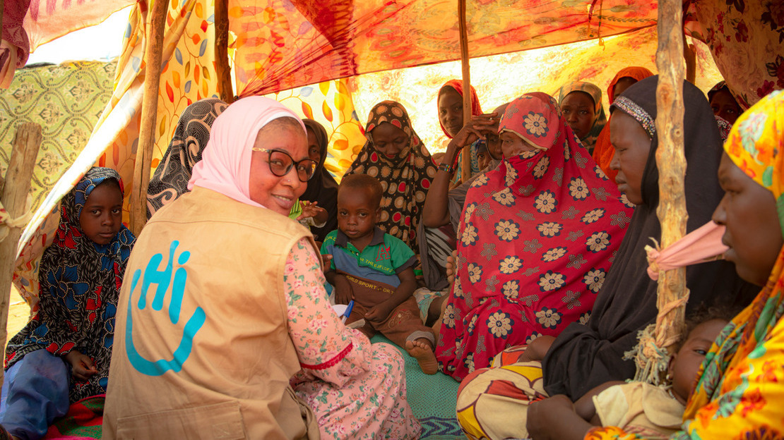 Women sit under a tent, chatting with an HI employee.