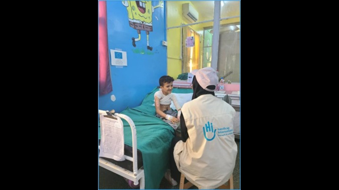 A child sits on a hospital bed covered with a green sheet in a colorful hospital room, facing a person wearing a vest with a logo. The room contains medical equipment, documents on the bed, and illustrated wall art.
