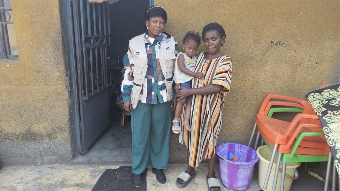 Two women, one holding a little girl in her arms, are standing in front of the half-open door of a house.