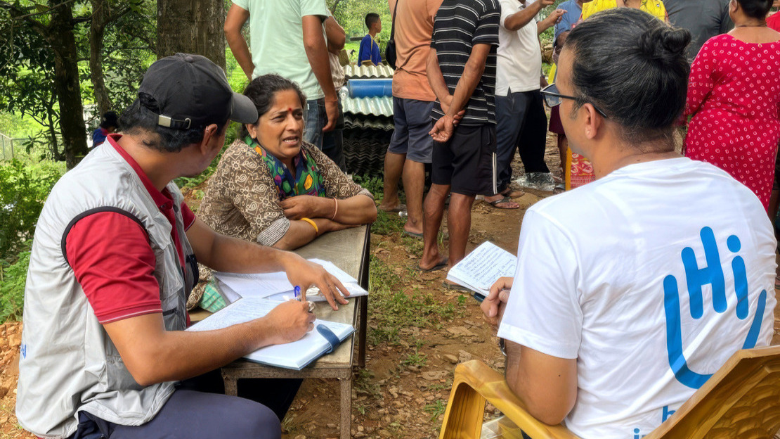 Two men, one wearing an HI shirt, are talking to a woman. Behind them, more people are standing and talking to each other. We can see a background of hilly vegetation.