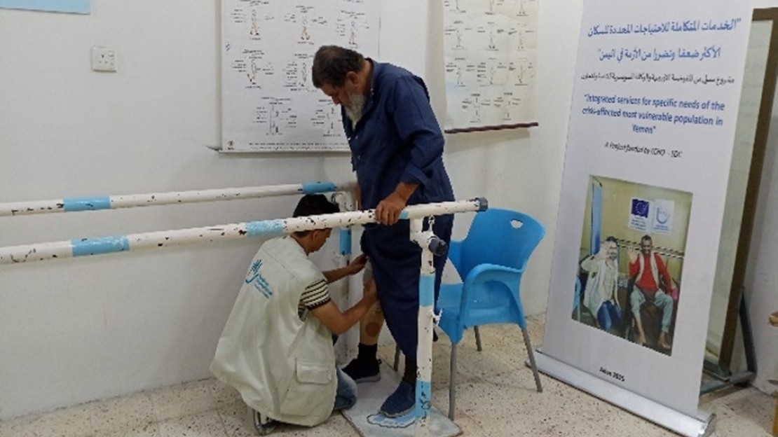 A person is standing between parallel rehabilitation bars in a room. Another person is fitting a prosthetic leg to the person’s lower limb. The room contains technical posters, a blue plastic chair, and an information panel on inclusion and rehabilitation.