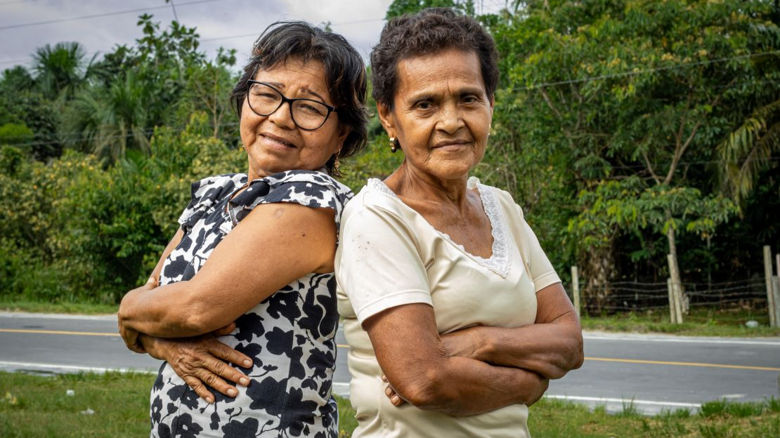 Portrait en gros plan de deux femmes âgées qui regardent la caméra en se faisant une accolade. Derrière elle, on devine une route bitumée et de la végétation.