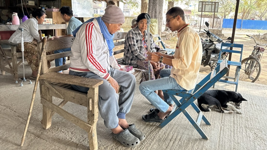 Three people are sitting on wooden benches under an open shelter. A younger person, sitting on a blue chair, is holding a small device and showing it to two people sitting on a bench across from them. In the background, other people are visible in a space that appears to be a workshop or a small shop. A dog is lying on the ground near the scene. A motorcycle and a bicycle are parked on the right. The floor is concrete, and several objects, including a stick lying near a bench, are visible around the area.
