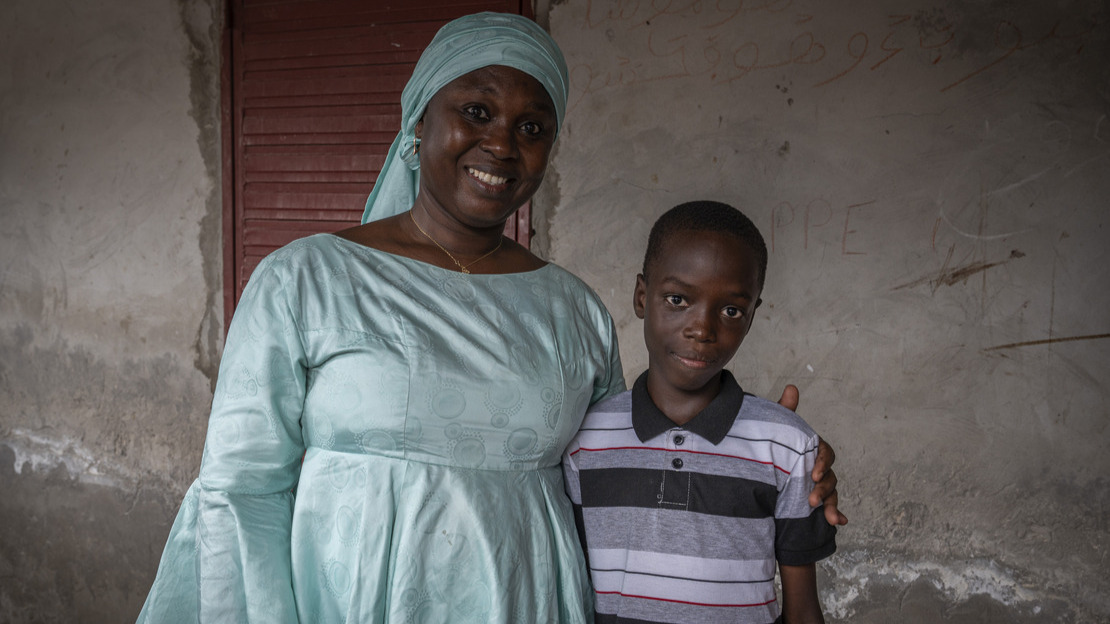 A close-up portrait of a woman holding a young boy by the shoulder. Both are looking at the camera and smiling.