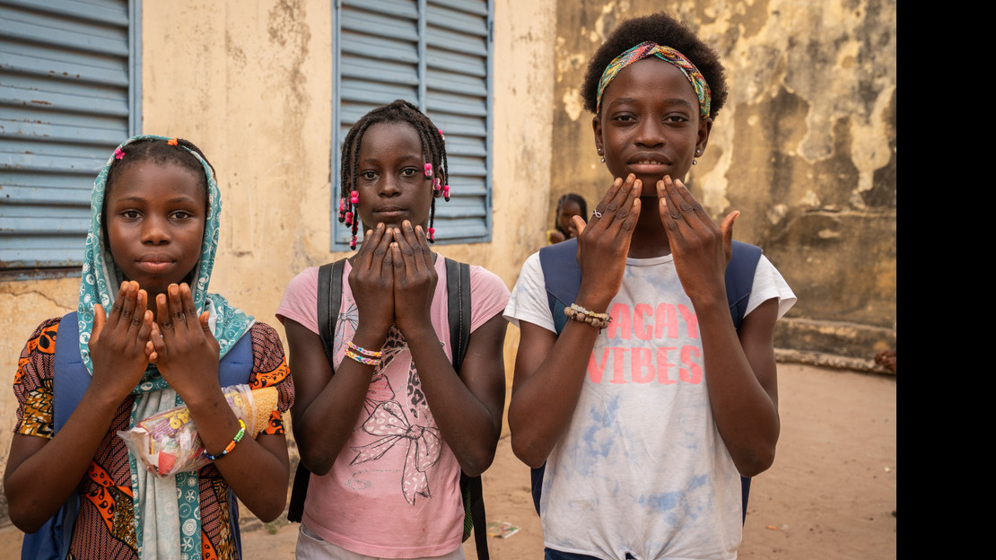 Three young girls are standing side by side, each with both hands held at the bottom of their faces, their open palms facing inwards.