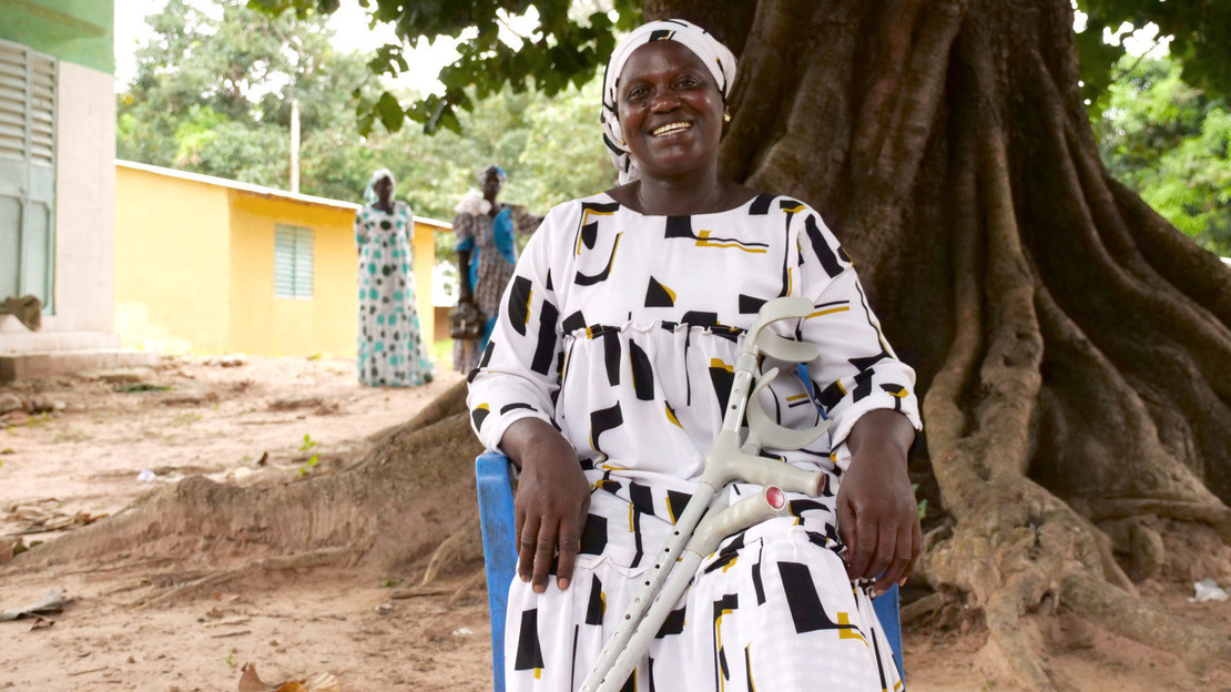 A woman sits on a blue plastic chair in front of a large cheese tree. Dressed in a white dress with black and yellow patterns, she smiles at the camera. Crutches rest on her legs.