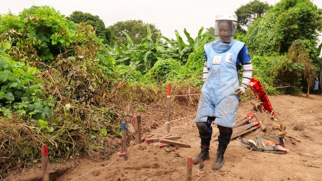 A woman in demining gear stands in front of a clearance lane marked by blue and red stakes, on a dirt road. Behind, lush vegetation.