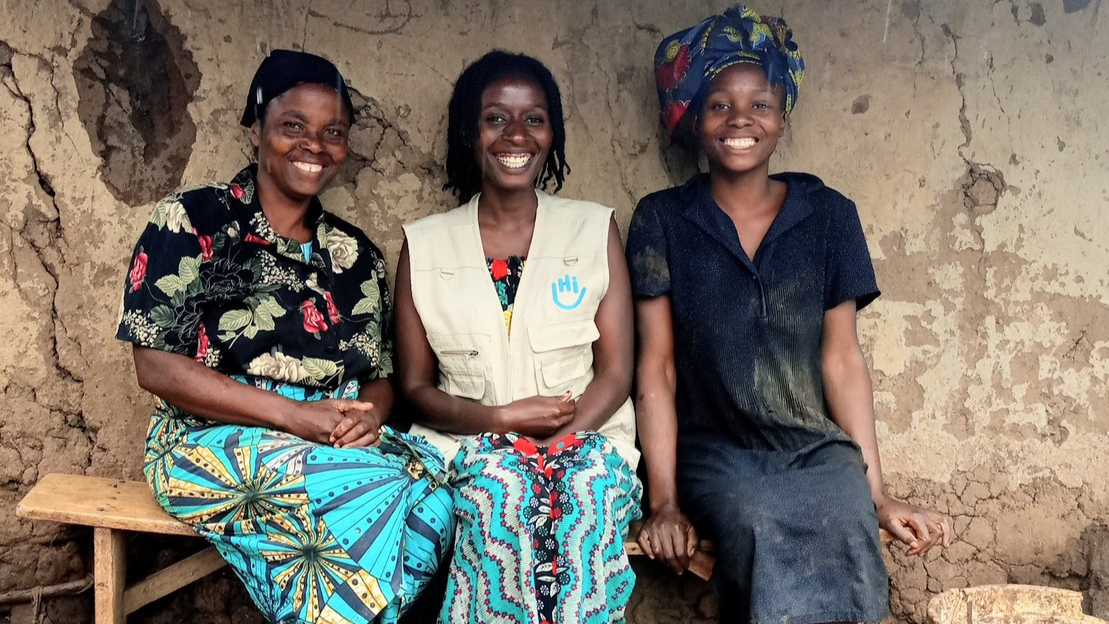 Beatrice, an HI mental health and psychosocial worker and Rose sit on a bench in front of their home. The three women are smiling.