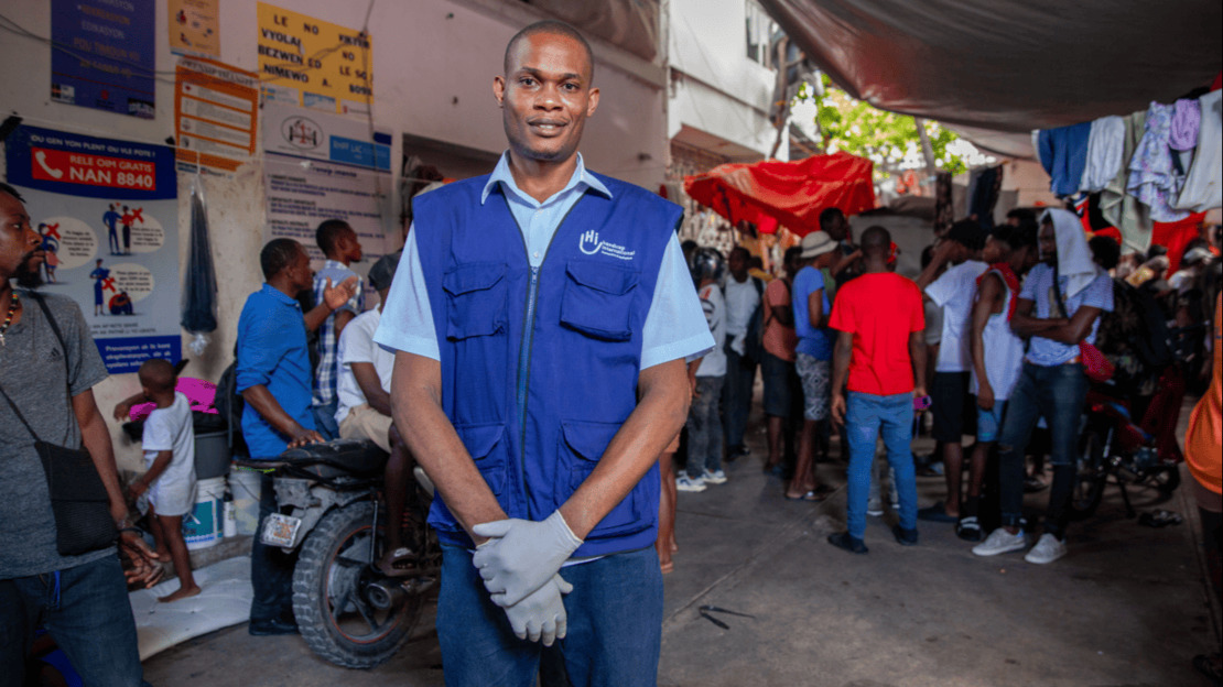 Portrait of a smiling man wearing work gloves and a HI jacket. Behind him, one can make out a room where a group of people are chatting and where laundry is drying.