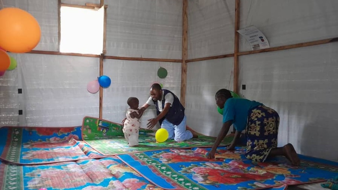 In a carpeted room, a man opens his arms to hold a baby. A woman crouches beside them. The walls are made of canvas, and balloons hang from them.