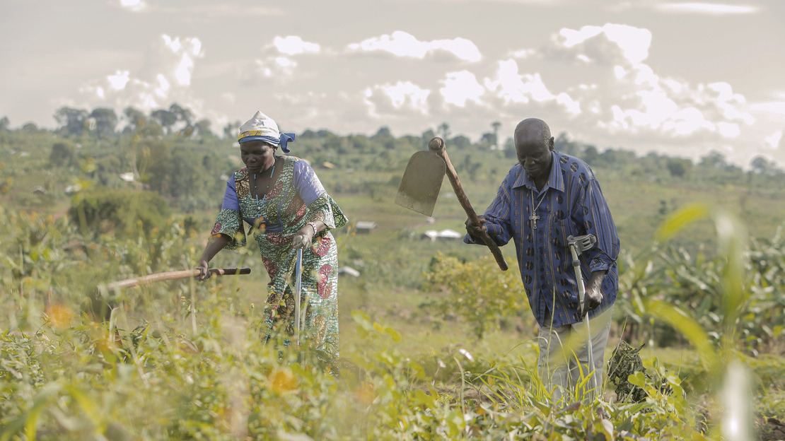 Two people, a woman and a man, are working in a field. Each is holding a spade in his right hand and a crutch in his left.