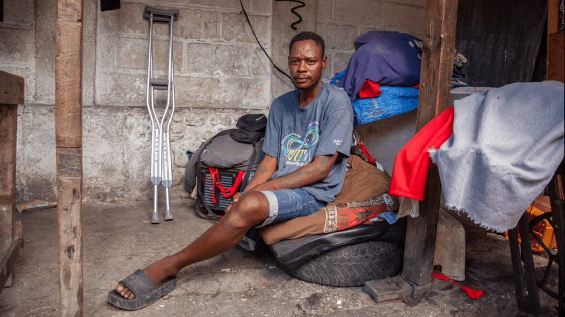 A one-legged man is sitting on a pile of belongings, in a space cluttered with fabrics, bags and other objects. Behind him, leaning against a brick wall, are two new crutches.
